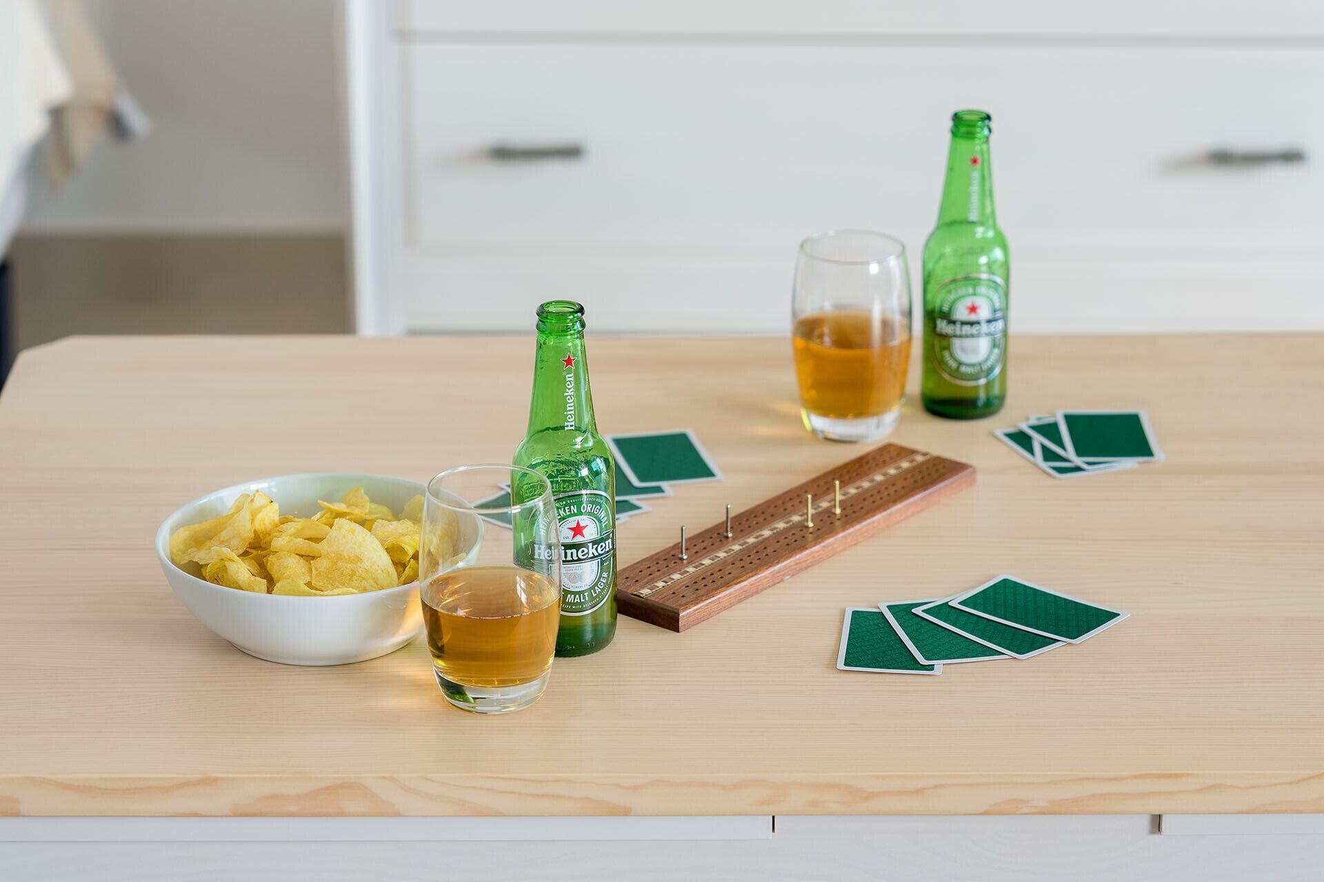 A wooden table with a bowl of chips, two glasses of beer, a pack of playing cards, and a wooden board game.