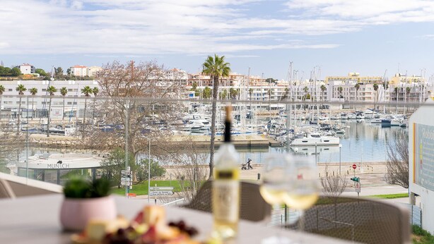 Une table à manger avec une bouteille de vin et une assiette de nourriture, donnant sur une marina où l’on peut voir des bateaux et des bâtiments.