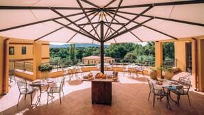An outdoor patio with multiple tables and chairs, a central wooden table, and a view of a building and trees in the background. - Hotel with swimming pool in Arbus (Arbus)