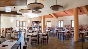 A spacious restaurant with multiple dining tables and chairs, a large kitchen area, and a high ceiling with exposed beams. - Hotel with swimming pool in Arbus (Arbus)