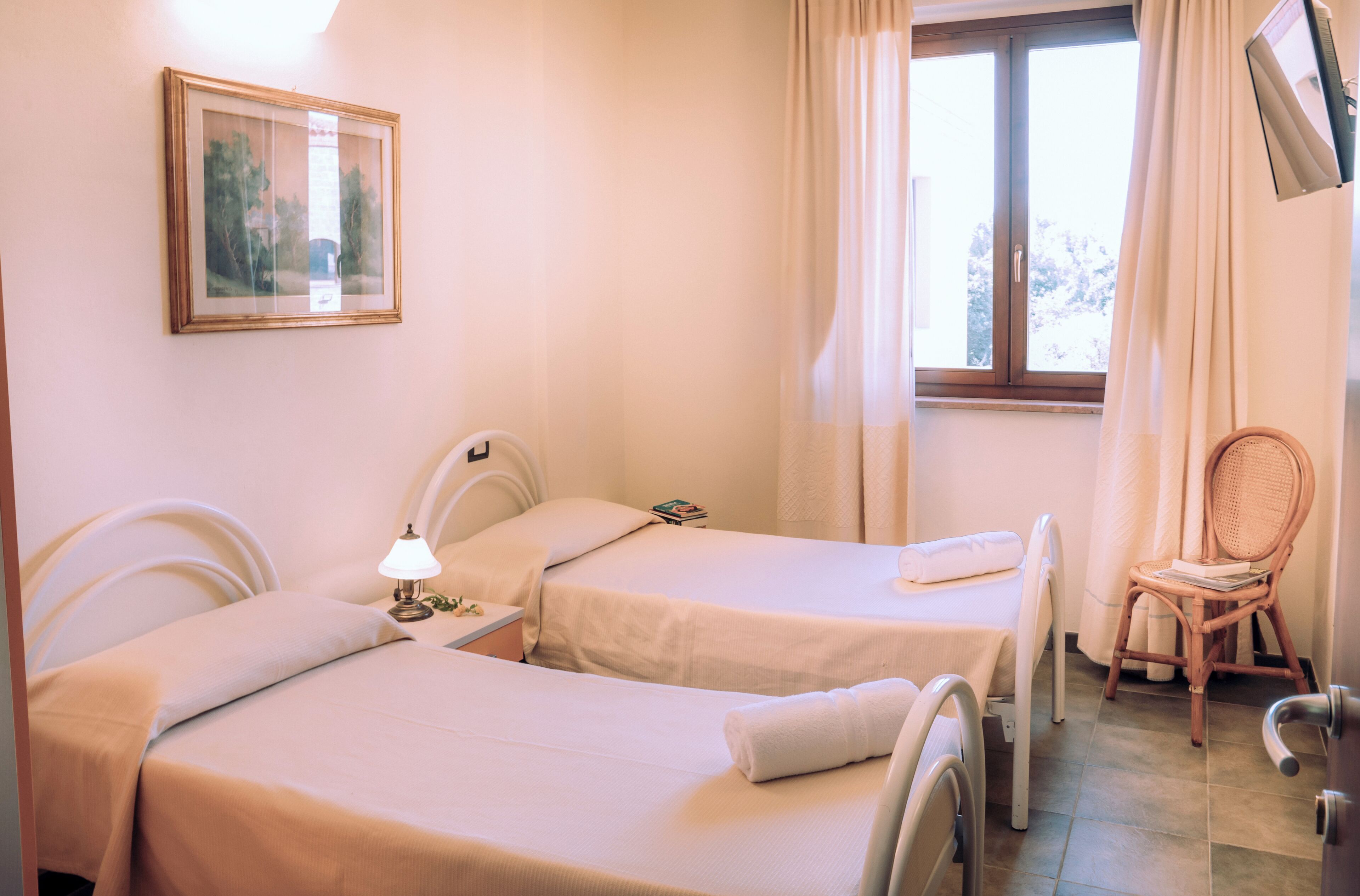 A bedroom with two beds, a bedside table with a lamp, a chair, and a window with curtains.