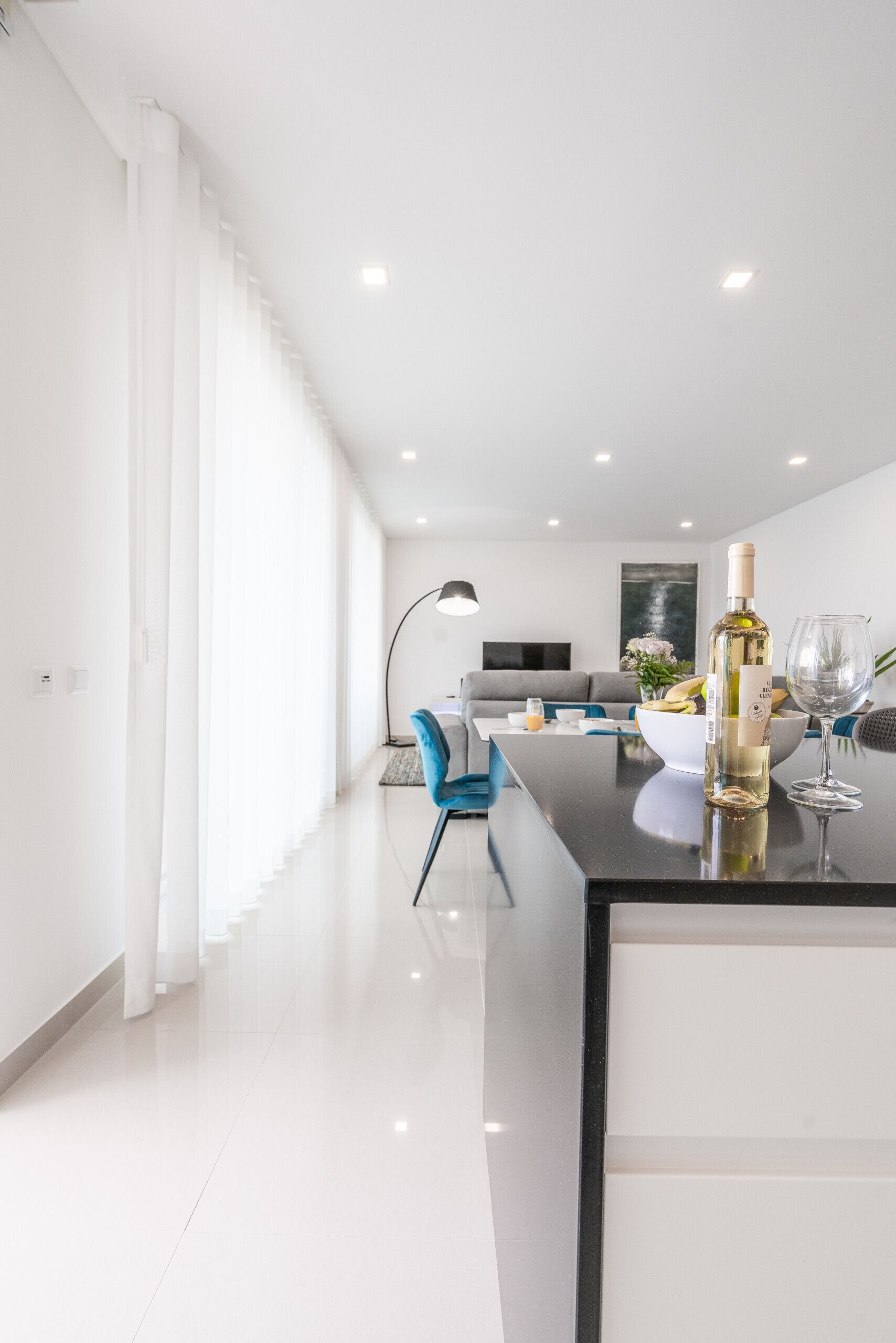 A modern living room with a dining area, featuring a kitchen island with a wine bottle and glass, a grey sofa, and a flat-screen TV.