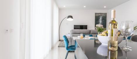 A modern living room with a dining area, featuring a kitchen island with a wine bottle and glass, a grey sofa, and a flat-screen TV.
