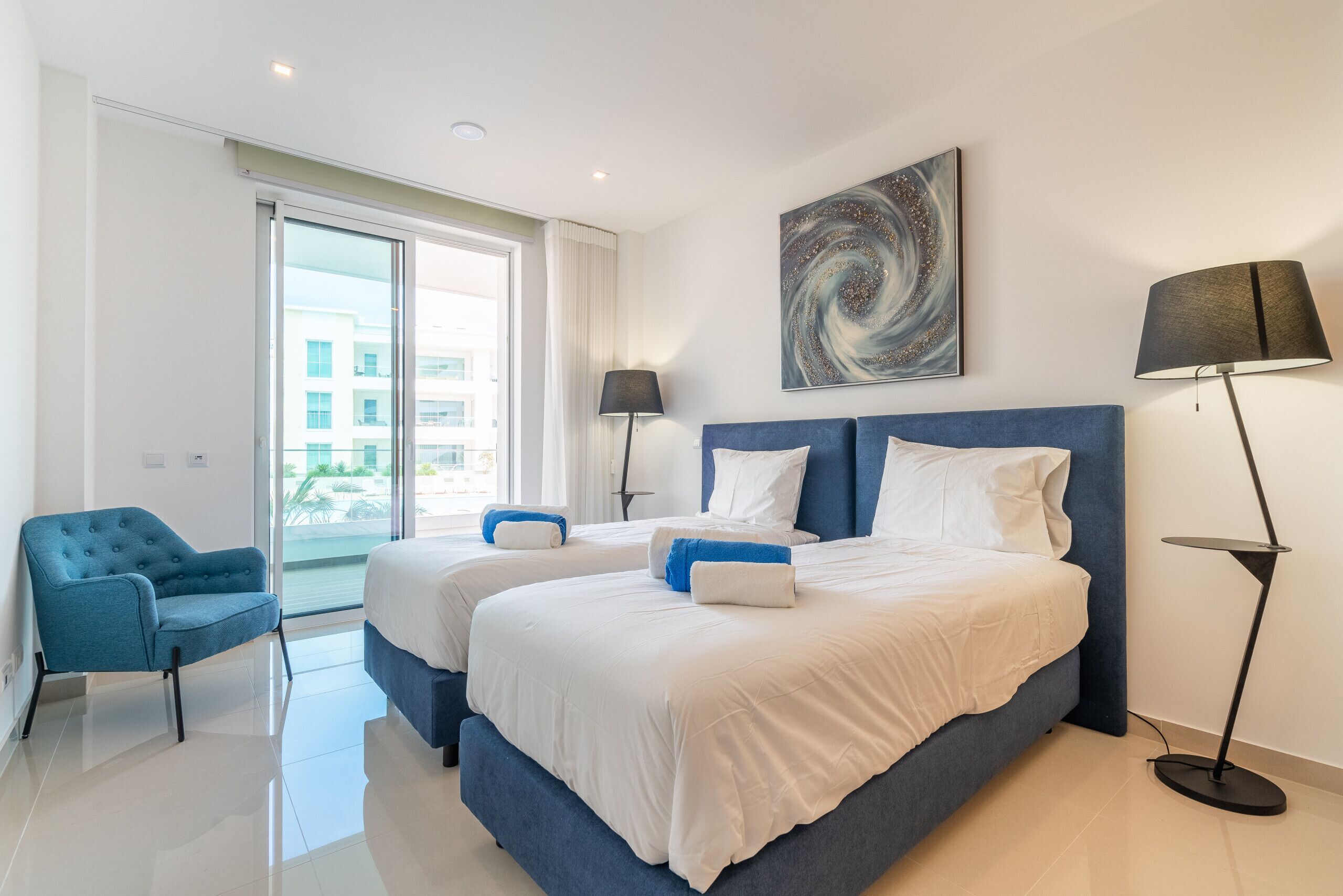 Habitación de hotel con dos camas, un sillón azul, una lámpara de pie y un cuadro abstracto grande en la pared.