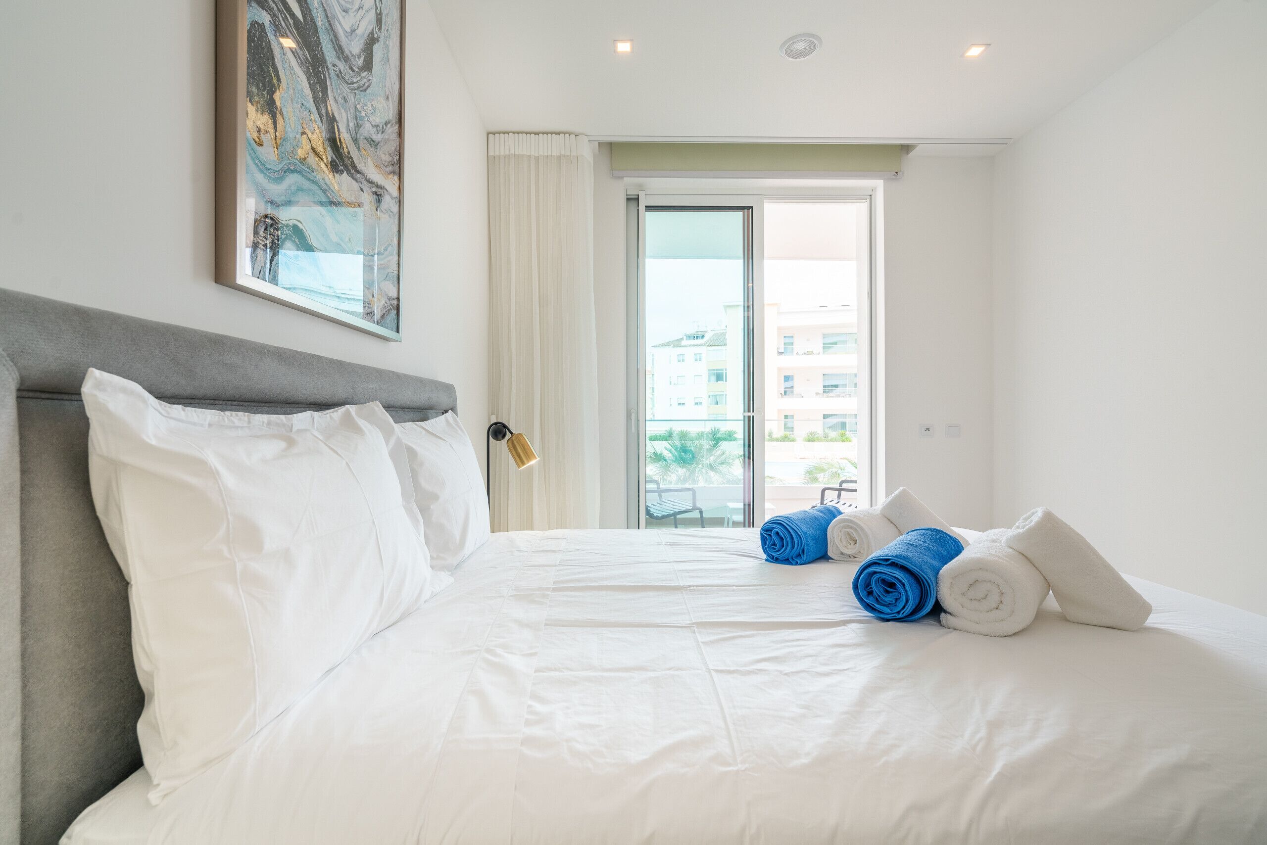 A hotel room with a large bed, white linens, a grey headboard, a painting on the wall, and a view of buildings through the window.