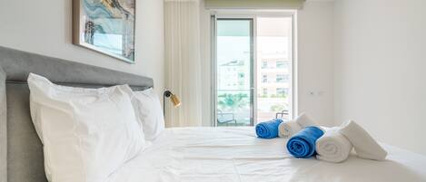 A hotel room with a large bed, white linens, a grey headboard, a painting on the wall, and a view of buildings through the window.