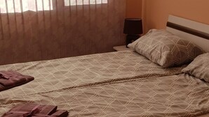 Una cama individual con una colcha de estampado geométrico y dos almohadas, una mesita de noche con una lámpara, y una ventana con cortinas de gasa.