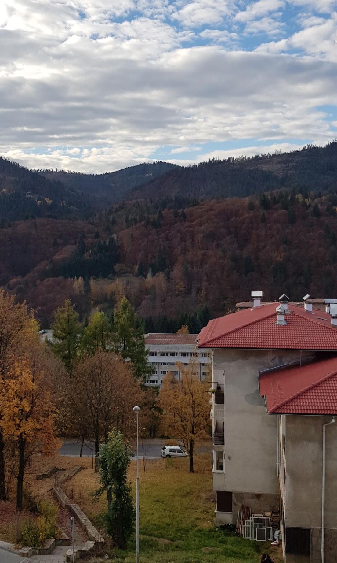 A building with a red roof, surrounded by autumn-colored trees and a hilly landscape in the background.