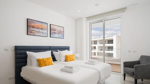 A modern hotel room with a large bed, two pillows, a chair, and a balcony with a view of buildings. - Lagos Marina View 1 With Pool by Homing (Lagos)