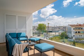 Un balcón con sofá y reposapiés, una botella de vino y vistas a un puerto deportivo y edificios.
