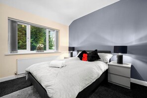 A bedroom with a bed, two bedside tables, a window with curtains, a radiator, and a lamp on the nightstand. - Harborne House, 4 bedrooms, Perfect for Families & Contractors (Birmingham)