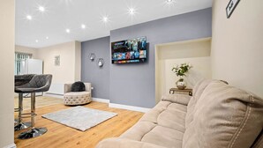 Un salon moderne avec un canapé gris, un pouf rond, un tabouret de bar gris et un téléviseur à écran plat fixé au mur, diffusant un film.