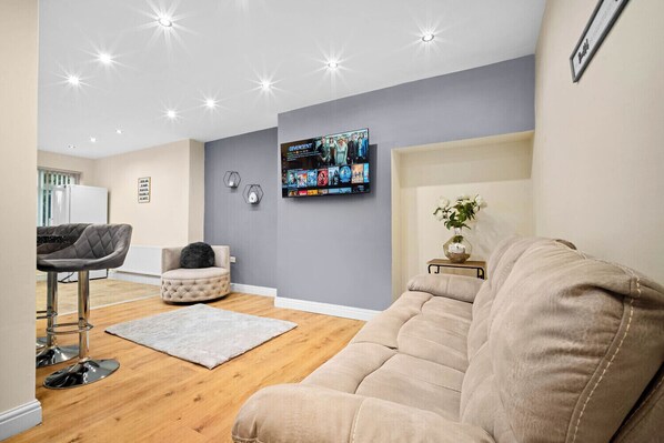 A modern living room with a grey sofa, a round ottoman, a grey bar stool, and a flat-screen TV mounted on the wall displaying a movie. - Harborne House, 4 bedrooms, Perfect for Families & Contractors (Birmingham)