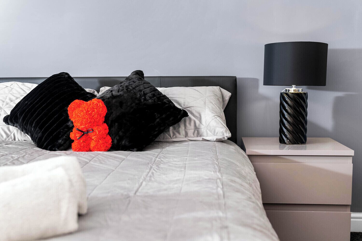 Een net opgemaakt bed met een grijze deken, zwart-witte kussens en een roos als decoratie. Op het nachtkastje staat een zwarte lamp en het nachtkastje is wit.
