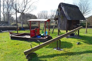 A playground with a seesaw, a red play structure, and a thatched-roof building.