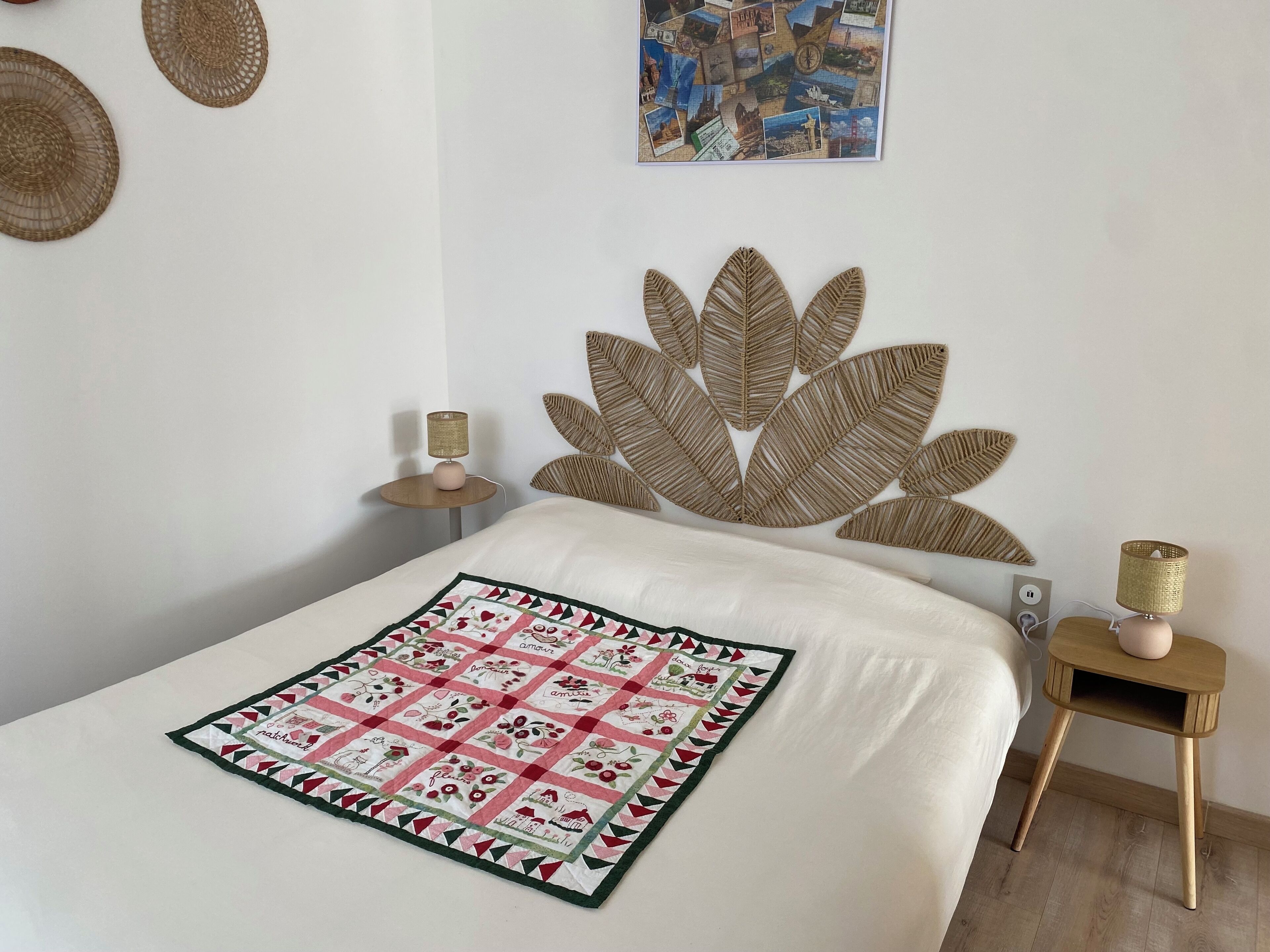 Een netjes opgemaakt bed met een decoratieve quilt, een wandmontage van een geweven hoofdbord, een klein nachtkastje met een lamp, en een ingelijst schilderij aan de muur.