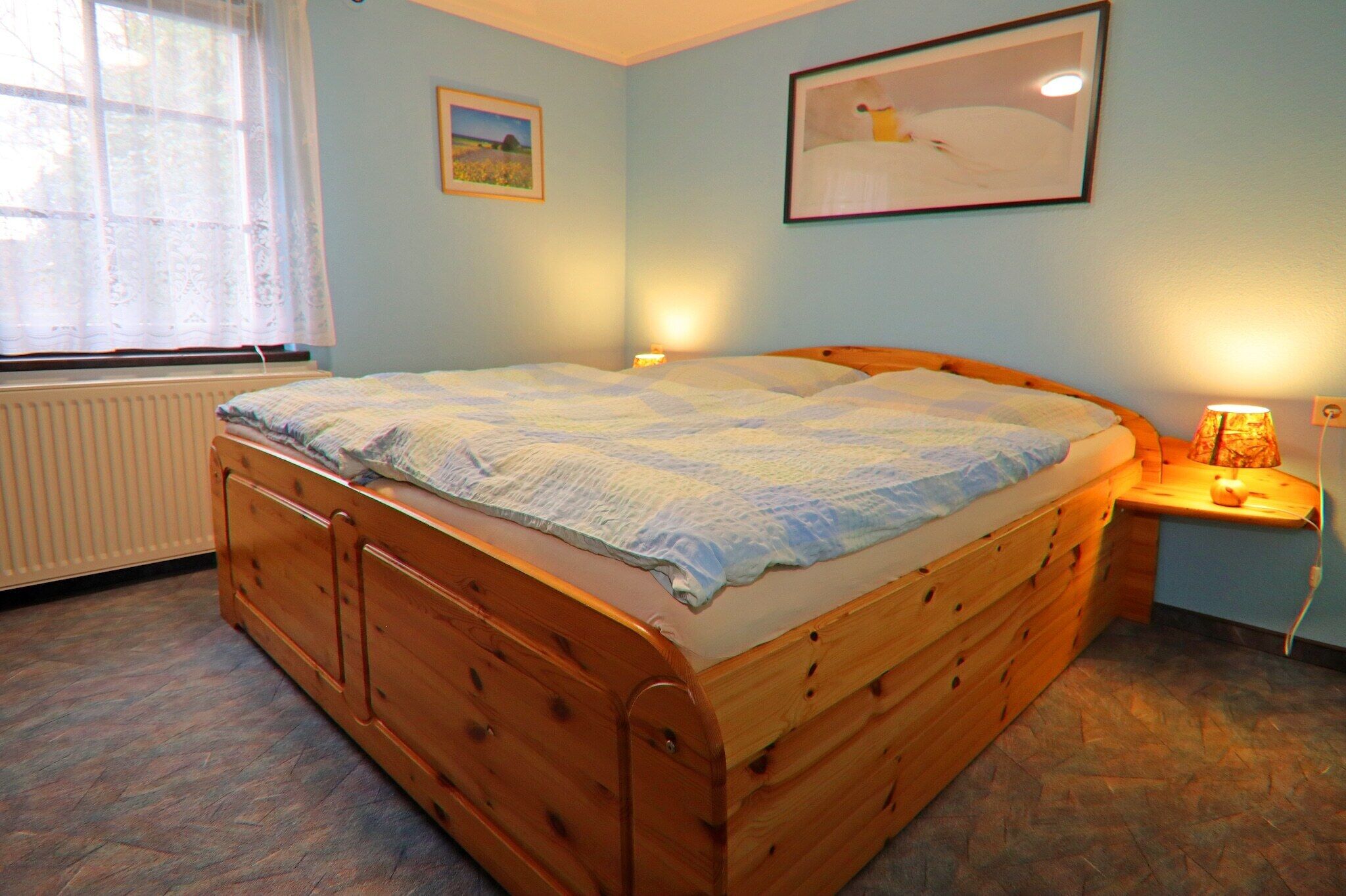 A bedroom with a wooden bed, a nightstand with a lamp, a window with a sheer curtain, and two framed pictures on the wall.