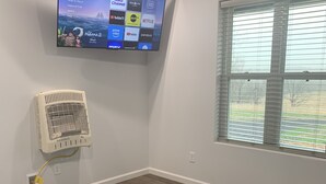 Ein Wohnzimmer mit einem an der Wand befestigten Flachbildfernseher, einem grauen, flauschigen Teppich, einem dunklen Sofa mit Zierkissen und einem Fenster mit Jalousien.