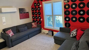 Un salón con sofá gris, pared de acento roja con discos de vinilo, una ventana grande y una lámpara en el techo.