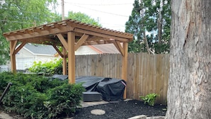 Una pérgola de madera con una zona de estar cubierta, rodeada por un jardín con mantillo y piedras para el paso.