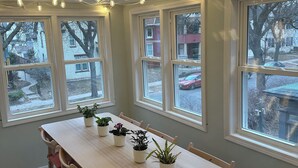 Una habitación con una larga mesa de madera, varias sillas, plantas en macetas y múltiples ventanas.