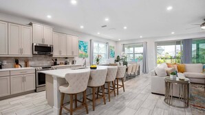 A modern kitchen with a central island, bar stools, and a dining area with a sofa and coffee table. - Luxury Retreat  7Bd Home - Close to Disney (Kissimmee)