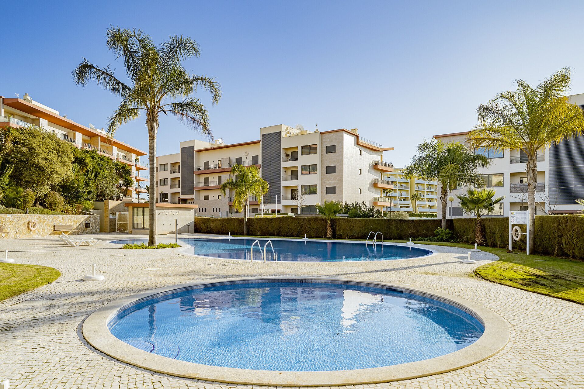 Una piscina rodeada por una zona pavimentada, con edificios de apartamentos modernos al fondo.