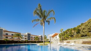 En swimmingpool med solstolar, palmer och moderna byggnader i bakgrunden.