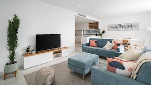 A modern living room with a flat-screen TV on a wooden stand, a blue sofa with cushions, a coffee table, and a potted plant. - Lagos Deluxe 1 With Pool by Homing (Lagos)
