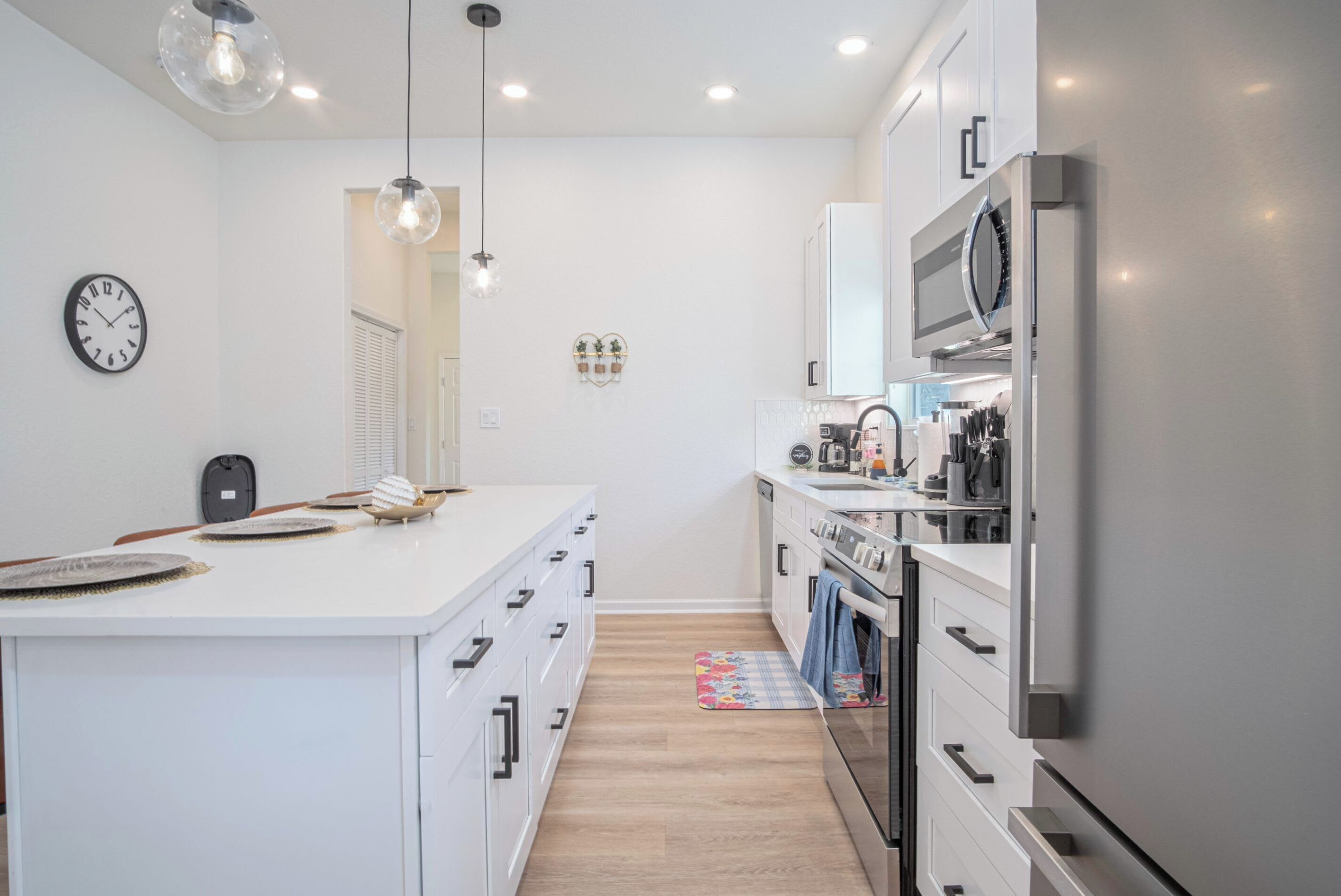A modern kitchen with white cabinetry, a central island, stainless steel appliances, pendant lighting, and a clock on the wall.