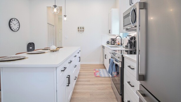 Une cuisine moderne avec des meubles blancs, un îlot central, des appareils en acier inoxydable, des luminaires suspendus et une horloge au mur.