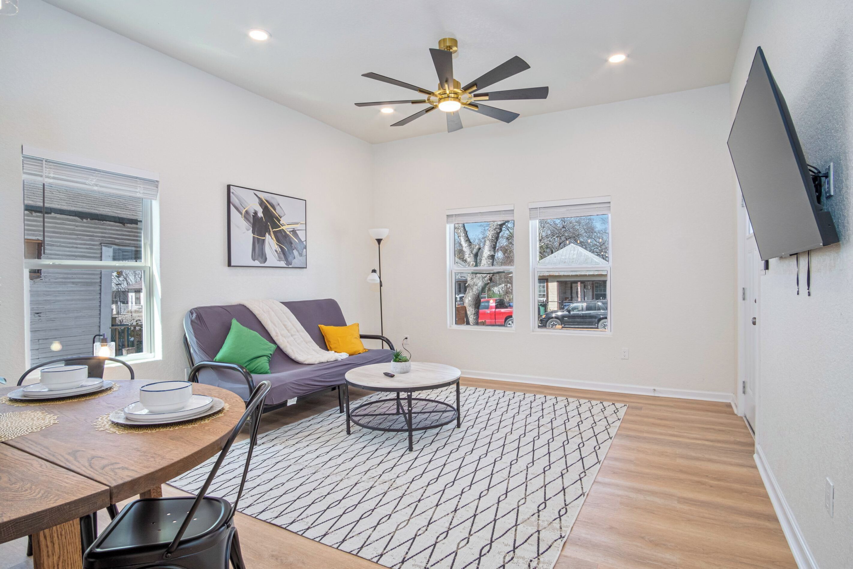A modern living room with a round dining table, a sofa with cushions, a ceiling fan, and a mounted TV.
