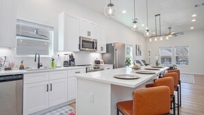 A modern kitchen with white cabinets, stainless steel appliances, a central island with bar stools, pendant lighting, and a view of the outside through large windows. - Feel, Chill and Enjoy 4br/3ba Near Fun Riverwalk! (San Antonio)