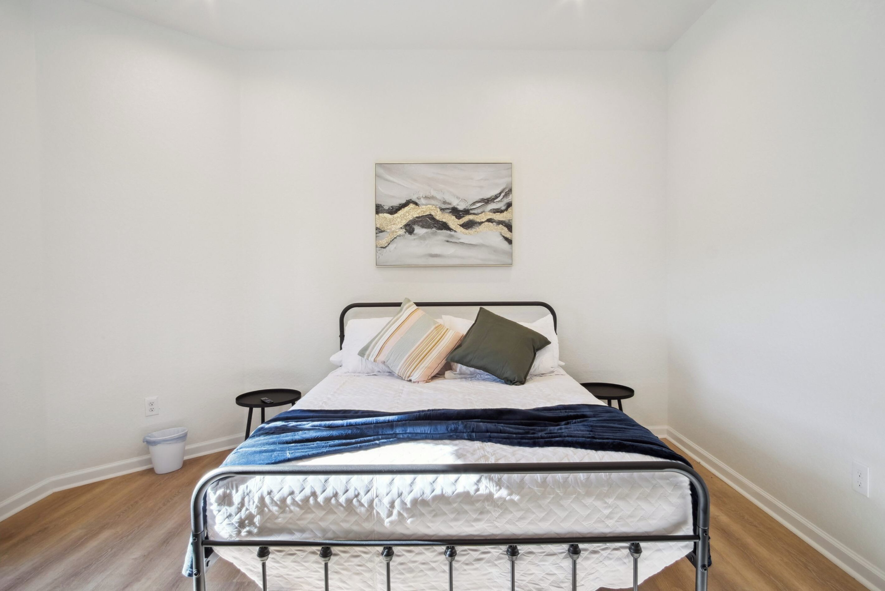 Een slaapkamer met een metalen bedframe, een witte geborduurde matras, een blauwe deken en een wanddecoratie met abstracte kunst.