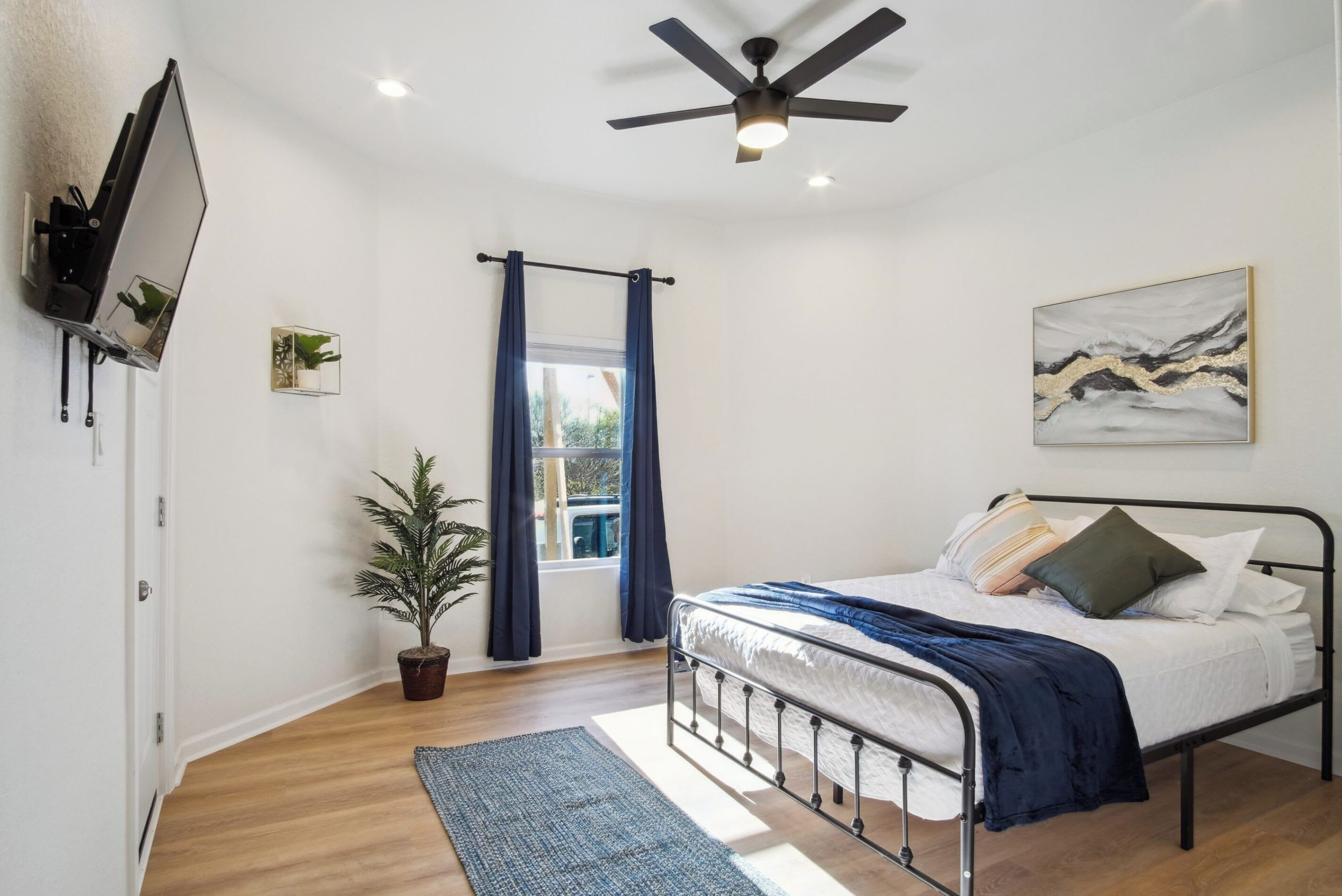 Quarto com cama, ventilador de teto, TV fixada na parede, janela com cortinas, planta em vaso e um quadro na parede.