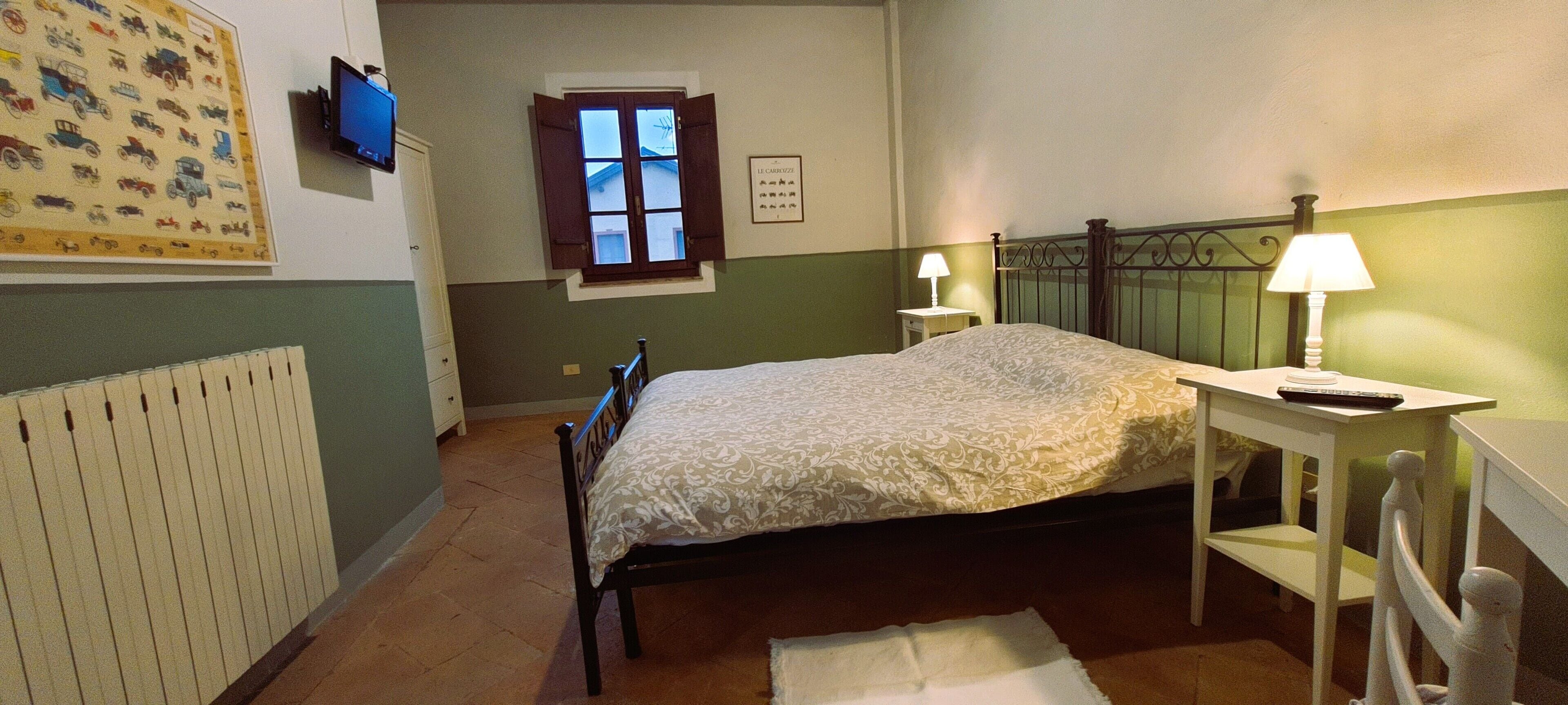 Un dormitorio con cama, una mesita de noche, una lámpara, una ventana y televisor montado en la pared.