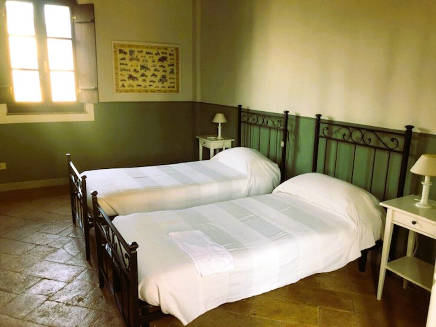 A room with two beds, a small table with a lamp, a wall-mounted picture, and a window with blinds.
