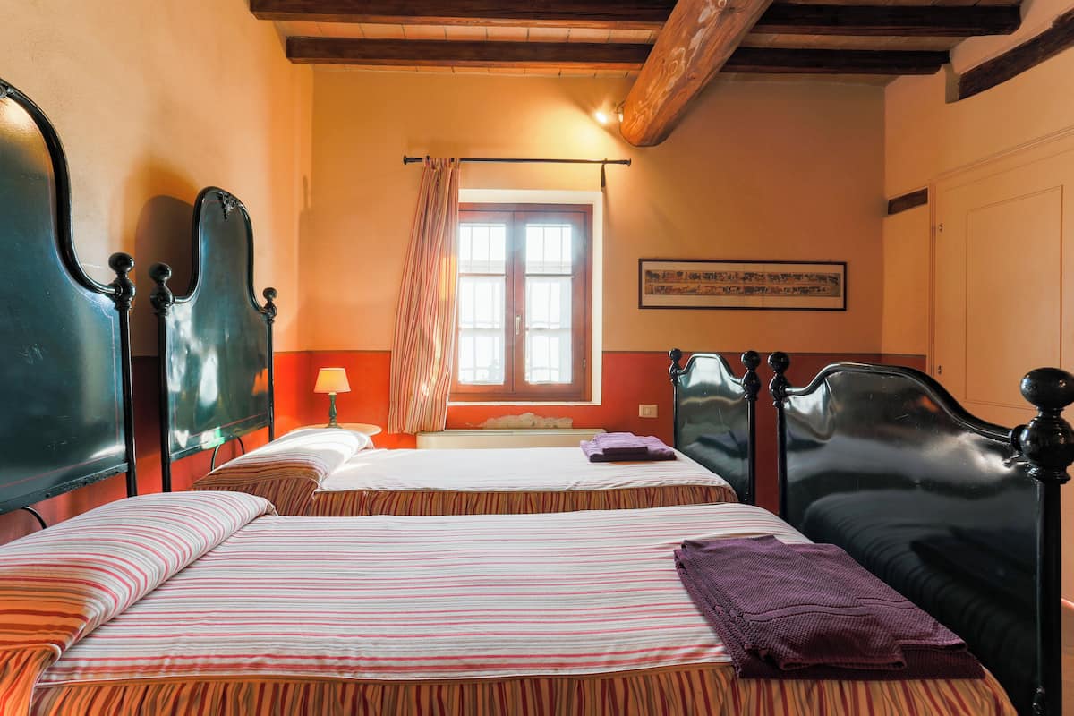 A room with two beds, a window with curtains, a framed picture on the wall, and a wooden ceiling beam.