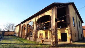 A building with multiple arches and a large open doorway, surrounded by a grassy area and a fence. - AGRITURISMO LE CORTI DELLA GUALDANA (Voghera)