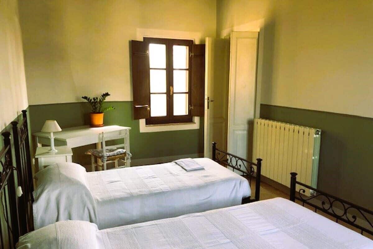 A room with two beds, a desk with a chair, a window with shutters, and a radiator.