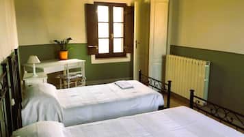 A room with two beds, a desk with a chair, a window with shutters, and a radiator.