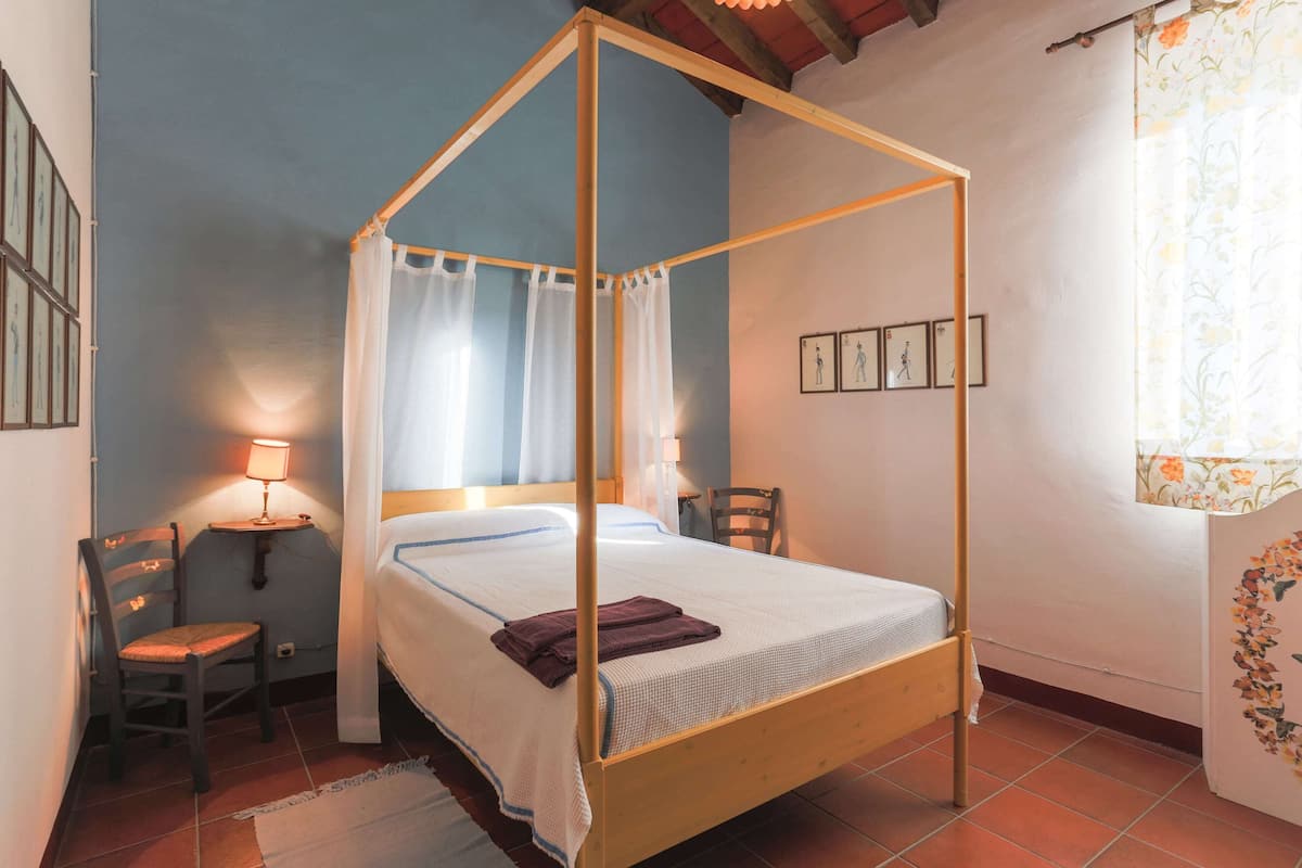 A four-poster bed with a canopy, a small bedside table with a lamp, a chair, and a window with curtains.