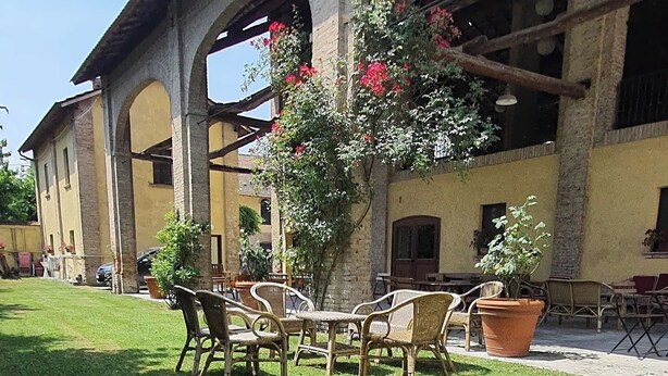 An outdoor seating area with wicker chairs and tables, surrounded by a well-maintained lawn and a building with arches and a stone facade. - AGRITURISMO LE CORTI DELLA GUALDANA (Voghera)
