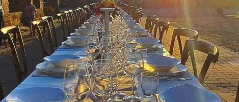 A long table set for a meal with chairs on both sides, under a clear sky with the sun low on the horizon.