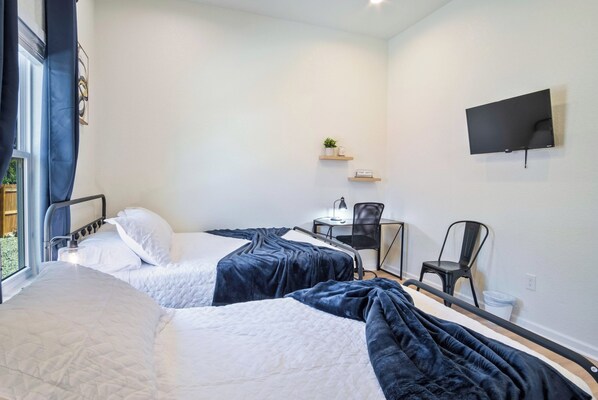 A hotel room with two beds, a desk, a chair, a TV, and a small shelf with a plant and a sign.