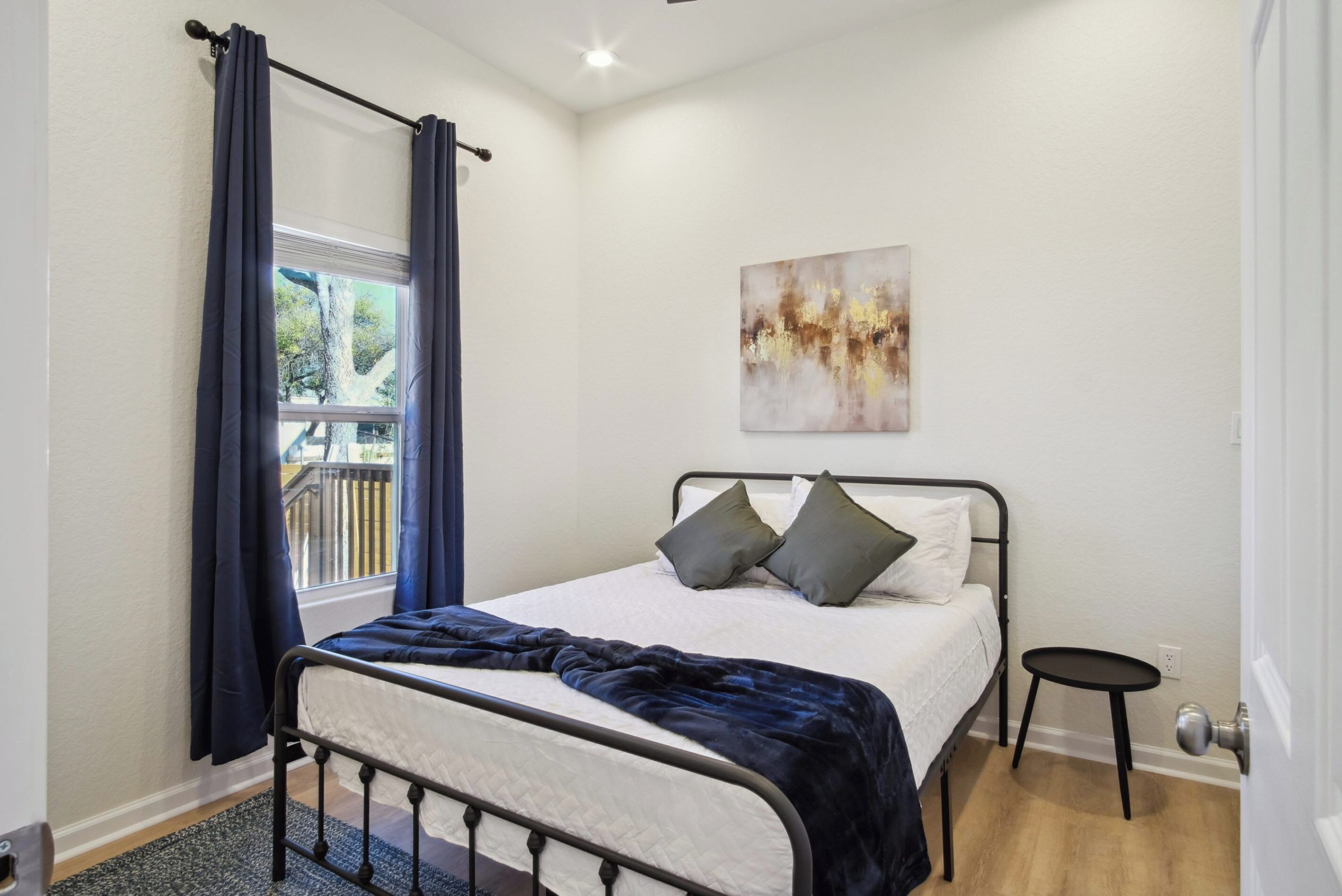 Un dormitorio con cama, ventana con cortinas, un taburete pequeño y una obra de arte colgada en la pared.