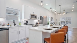 A modern kitchen with white cabinets, stainless steel appliances, a central island with bar stools, pendant lighting, and a view of the outside through large windows. - Feel, Chill and Enjoy 4BR/3BA Near Fun Riverwalk! (San Antonio)