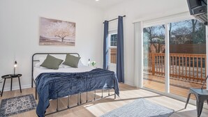 A bedroom with a bed, a painting, a window with curtains, a small table, and a view of a wooden deck. - Feel, Chill and Enjoy 4BR/3BA Near Fun Riverwalk! (San Antonio)