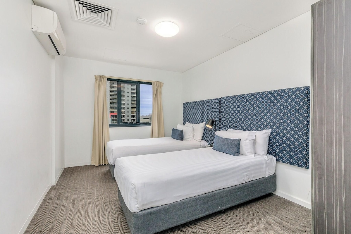 A hotel room with two beds, a patterned headboard, a window with curtains, and an air conditioning unit.