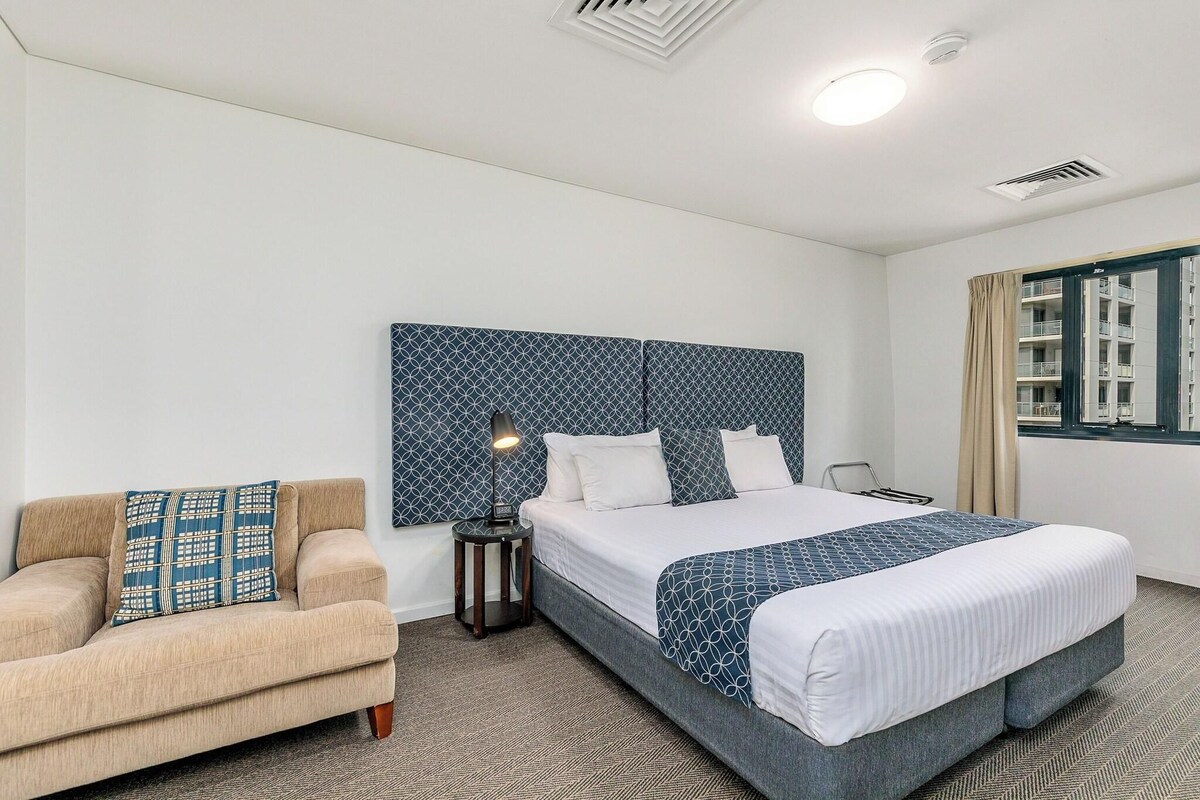 Une chambre d’hôtel avec un lit, une chaise, une table de chevet, une lampe et une fenêtre avec des rideaux.